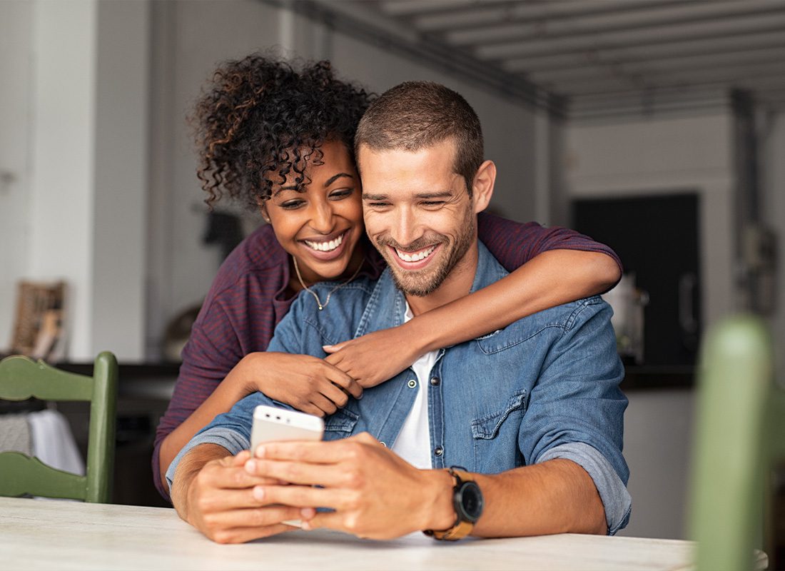 Read Our Reviews - A Young Couple Looking at a Phone While Smiling Together at Home